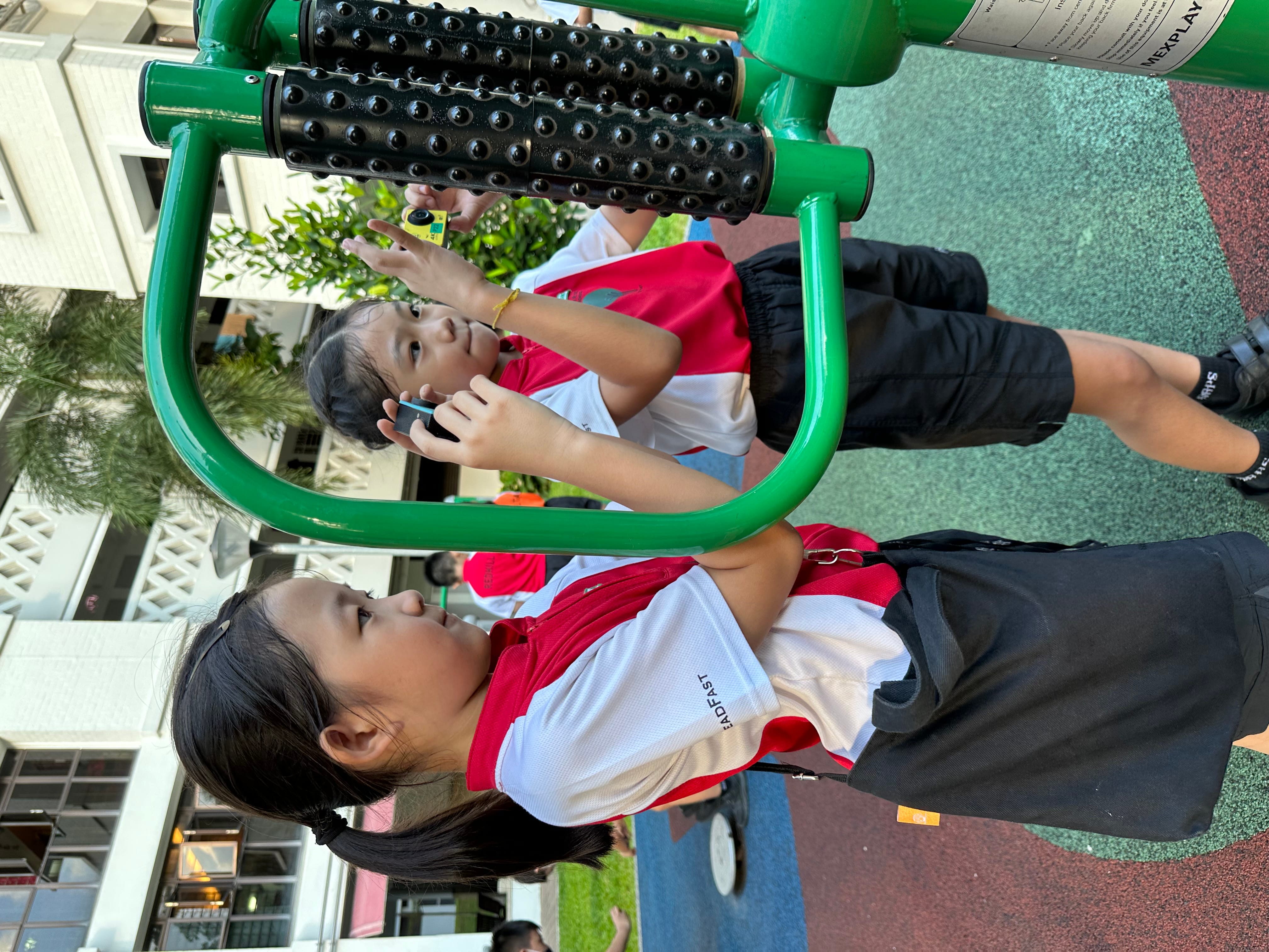 Two children in red and white school uniforms stand beside a green outdoor fitness machine with textured black rollers.